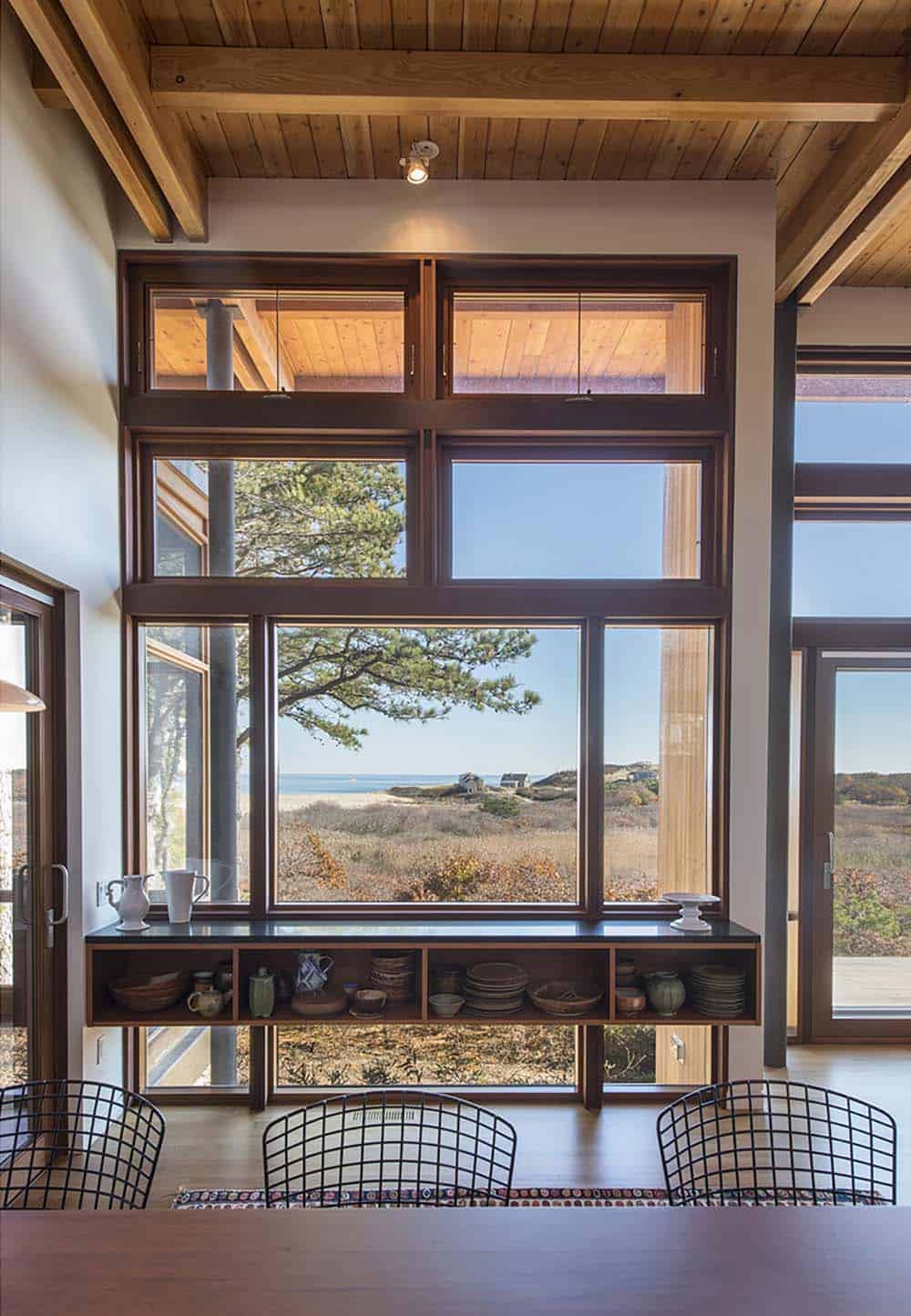 Tall wood-framed windows with a built-in open shelf below displaying pottery and dishes, framing a view of pitch pines, dunes, and ocean