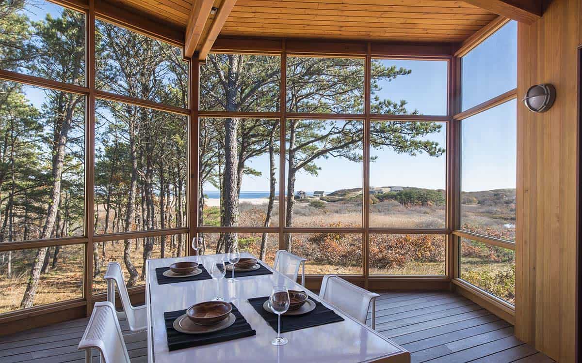Screened porch with wood-framed glass walls on three sides, a white dining table set for four, and a panoramic view of coastal pines, dunes, and ocean