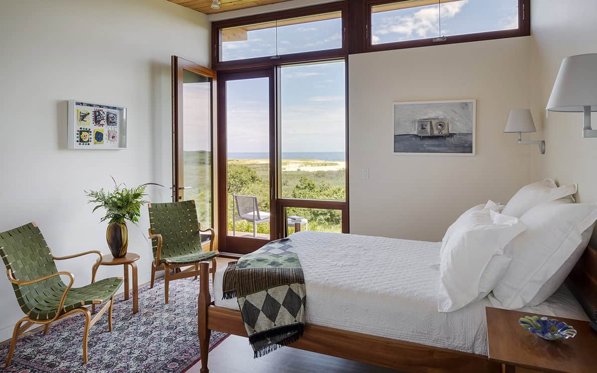 Bedroom with wood-framed corner windows opening to an ocean view, green woven lounge chairs, a wood bed frame with white bedding, and a Persian rug