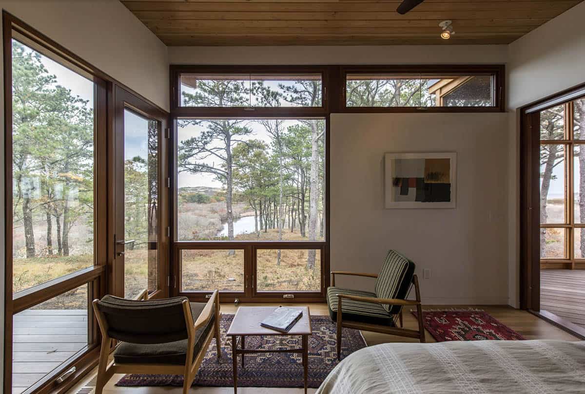 Bedroom sitting area with two mid-century wood-framed chairs, a low coffee table, and floor-to-ceiling windows overlooking coastal pines and a pond