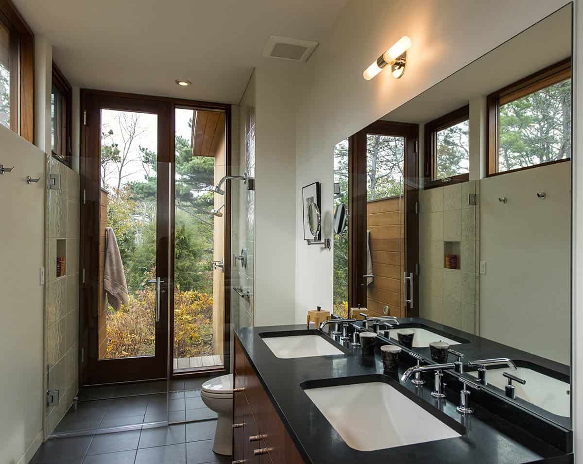 Primary bathroom with double undermount sinks set in a dark stone countertop, a full-wall mirror, glass-enclosed shower, and wood-framed windows with pine tree views