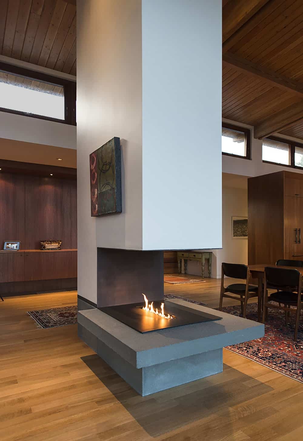 Double-sided fireplace on a raised bluestone hearth set into a white chimney column, with wood-paneled walls and exposed wood ceiling beyond
