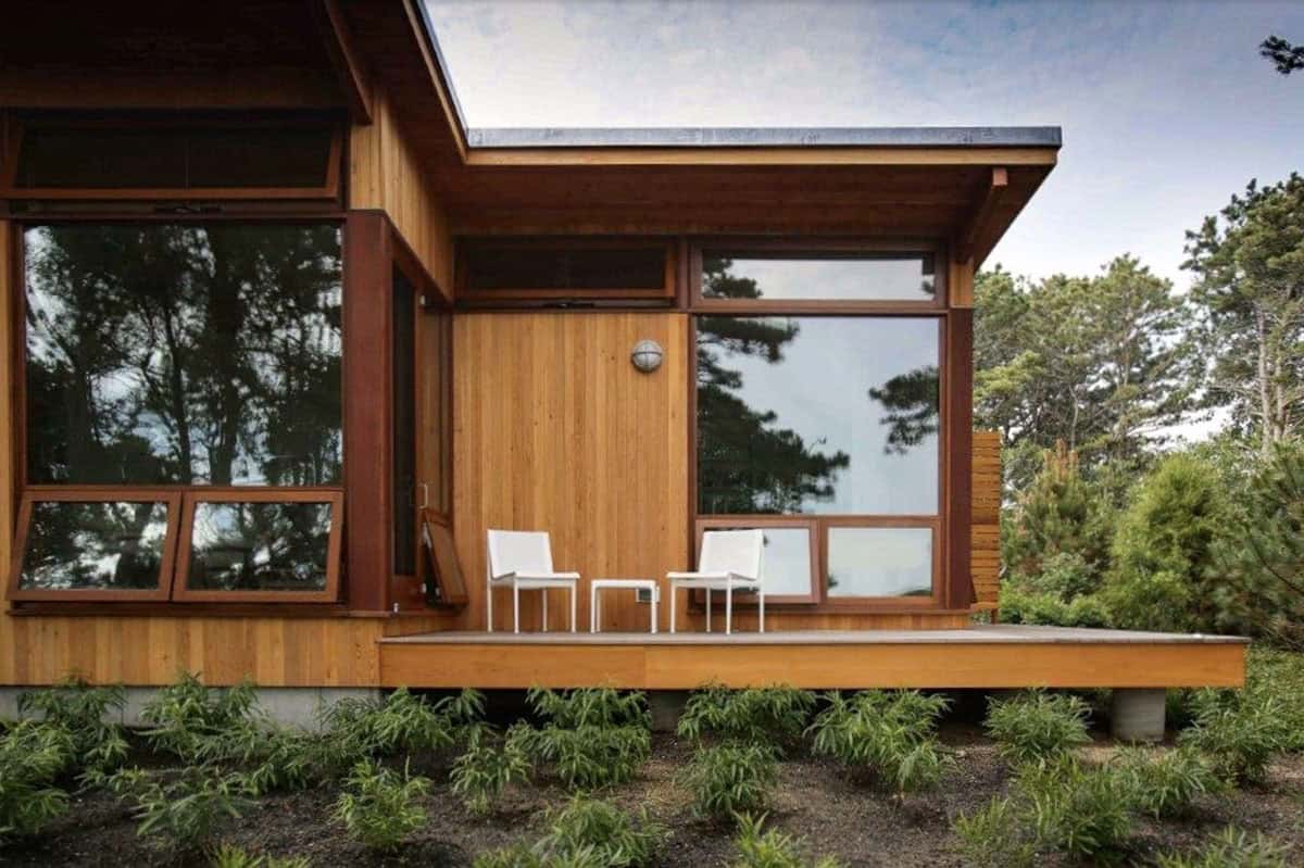 Cedar-sided bedroom wing exterior with a small wood deck, two white chairs, large wood-framed windows, and native plantings in the foreground