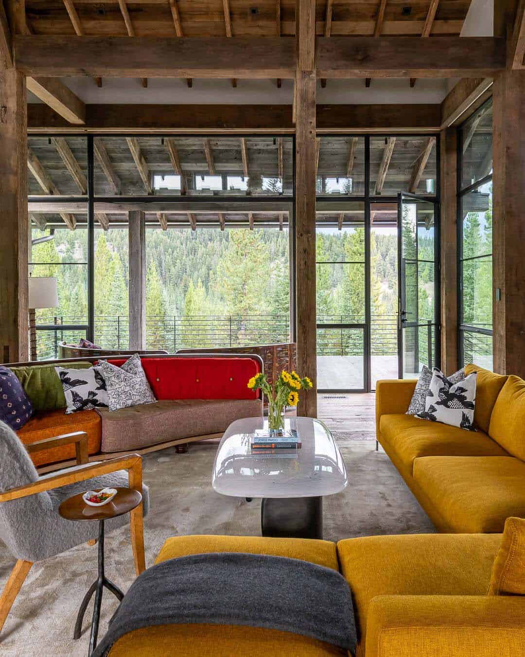 Living room with floor-to-ceiling steel windows, forest views, and mustard sofas
