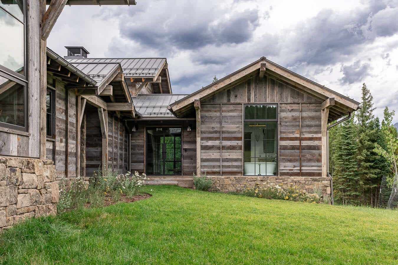 Rear exterior of reclaimed wood home with metal roof, stone foundation, and mountain meadow