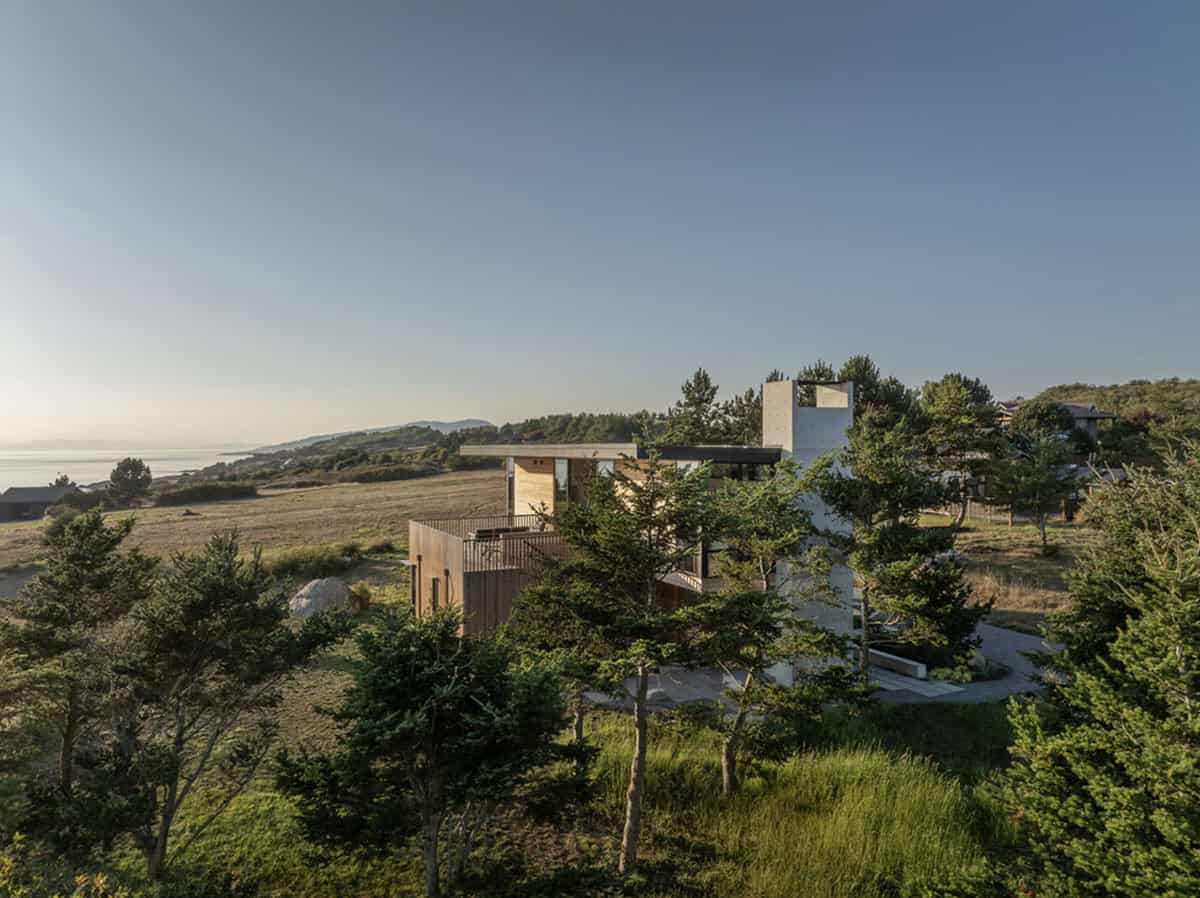 Aerial view of a modern beach house nestled among pine trees and coastal grassland, with the San Juan Islands shoreline visible in the distance