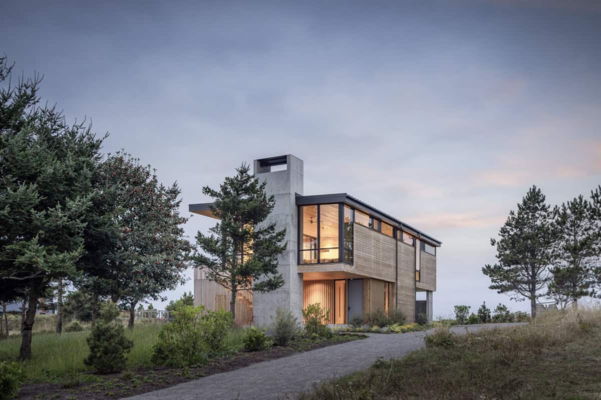 Twilight exterior of a modern beach house with a tall concrete chimney, cedar siding, and glowing interior lights