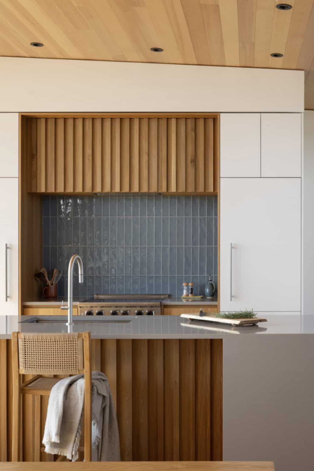 Kitchen nook with fluted wood range hood, blue tile backsplash, and a woven counter stool