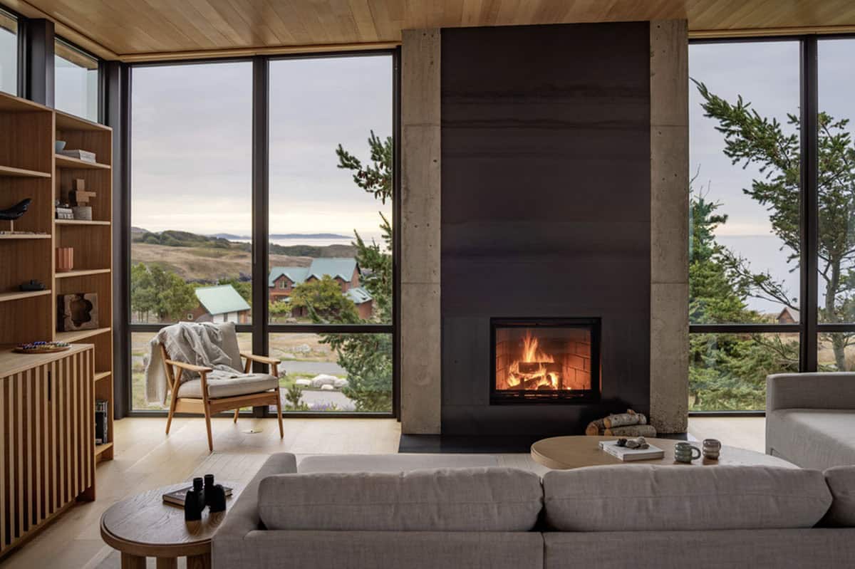 Living room with a tall concrete-and-steel fireplace, gray sofa, and panoramic windows overlooking the coastline
