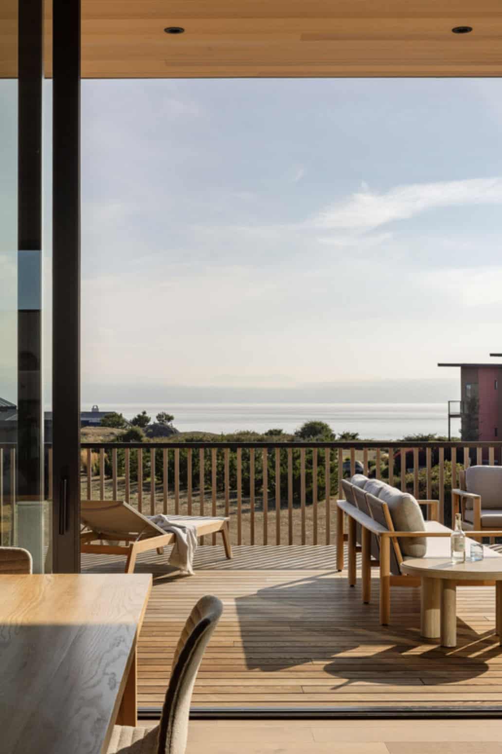 View from the dining area through open glass doors to a wood deck with lounge seating and ocean views