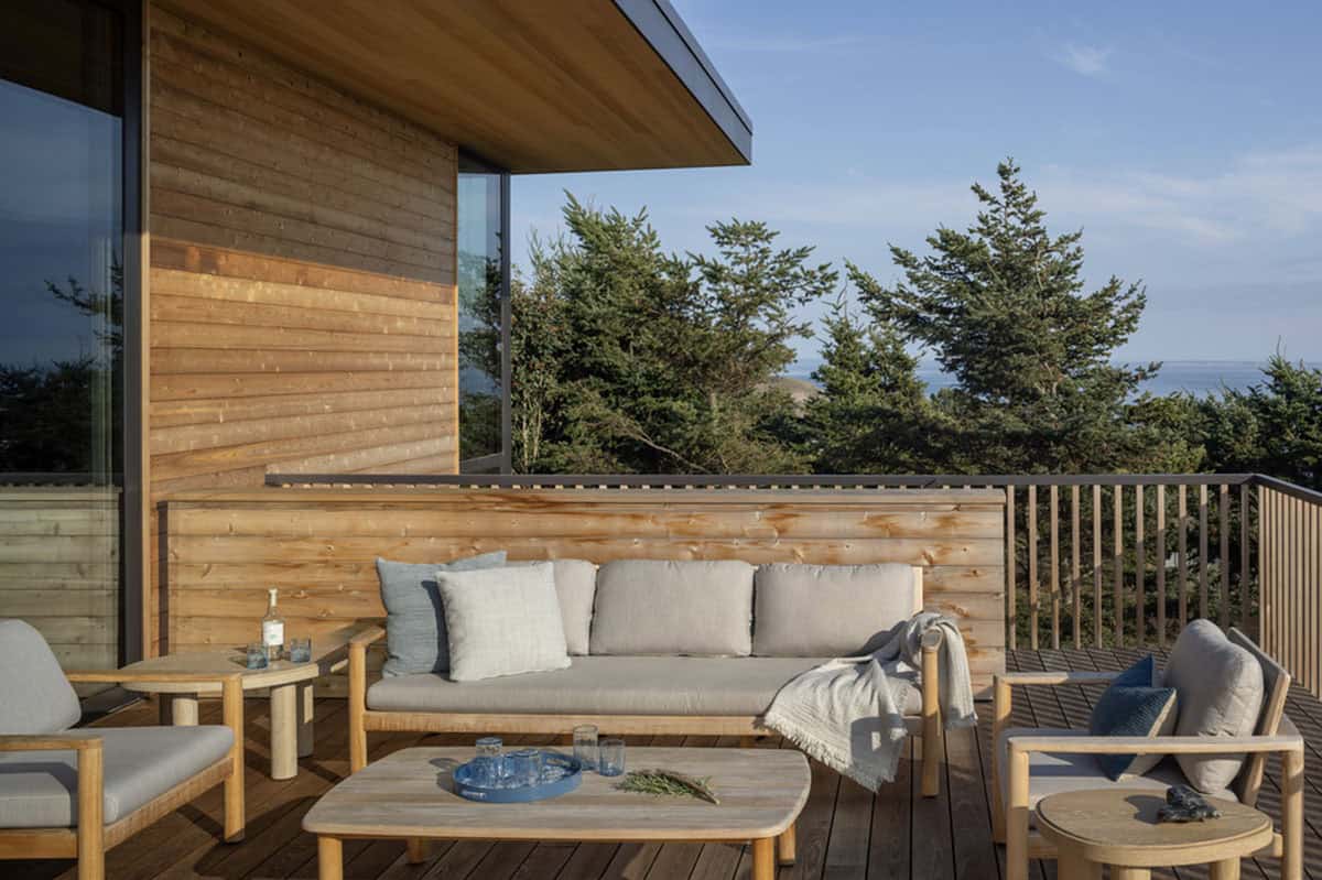 Outdoor deck lounge area with cushioned wood furniture, surrounded by pine trees and a glimpse of the sea