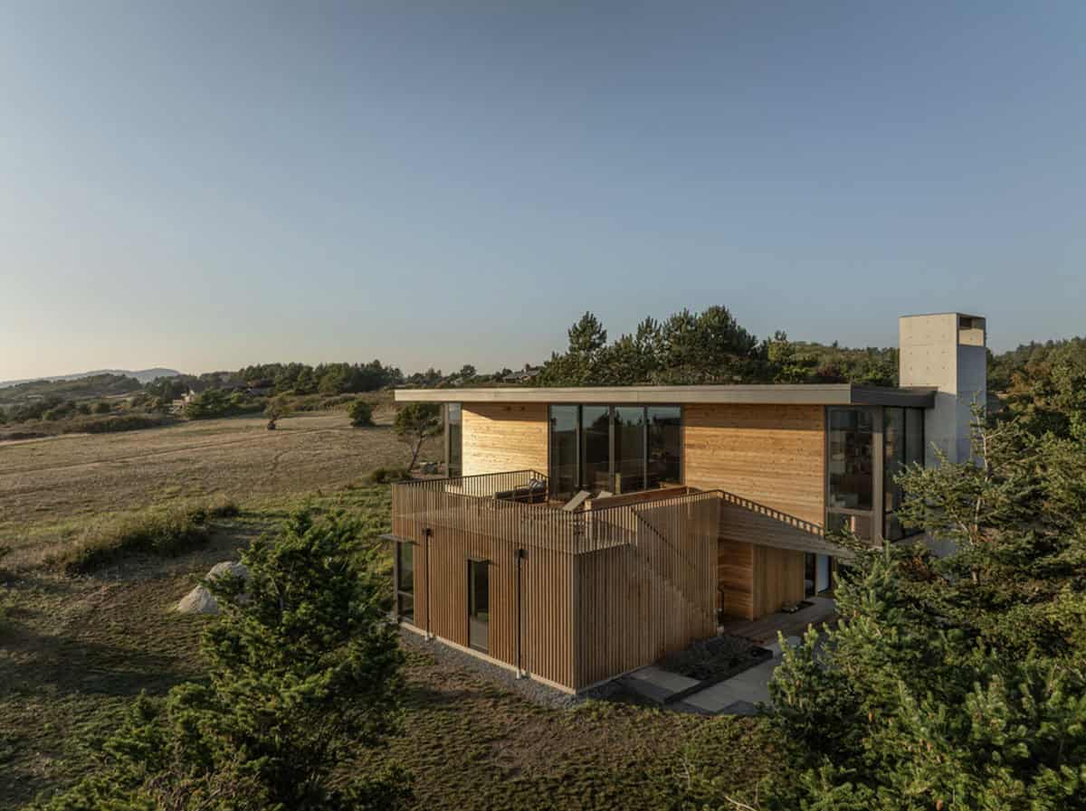 Exterior view of the modern beach house showing its cedar slat facade, concrete chimney, and surrounding wild grasses
