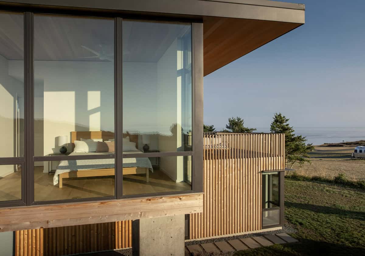 Exterior view into a glass-walled bedroom with a wood bed frame, set above a cedar-slatted lower level