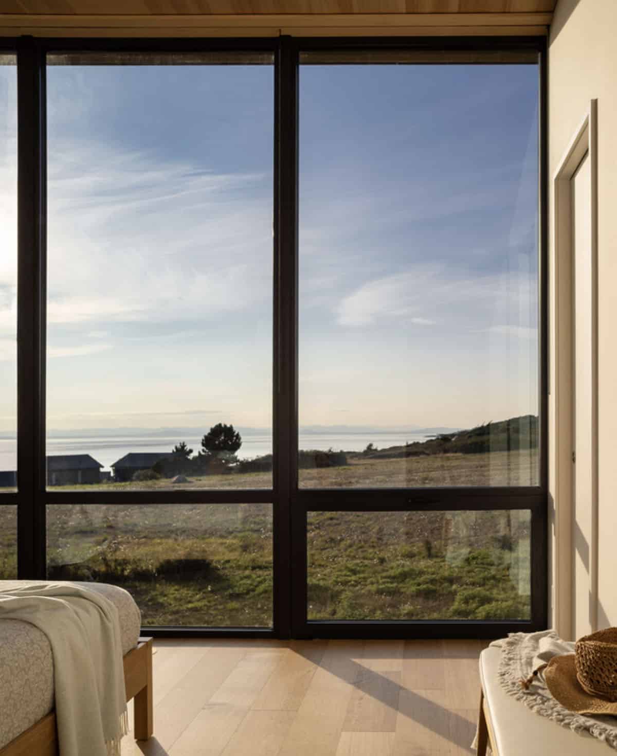 Bedroom view through floor-to-ceiling windows overlooking grassy fields and the distant shoreline