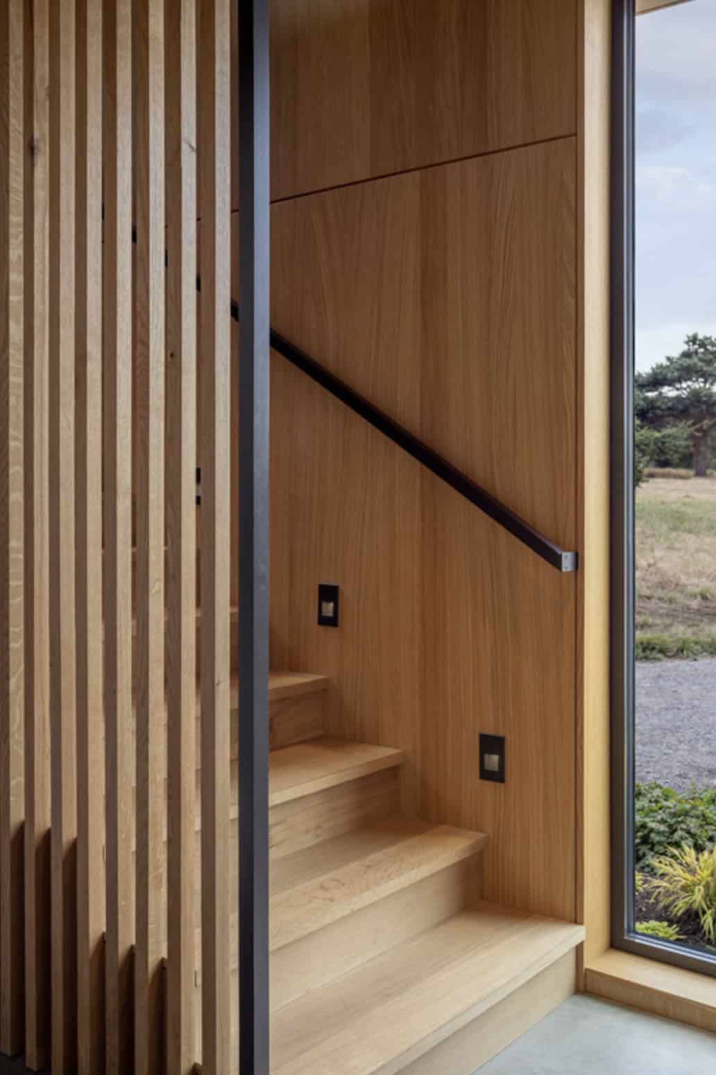 Interior staircase detail with oak paneling, a black handrail, and cedar slat screen beside a window