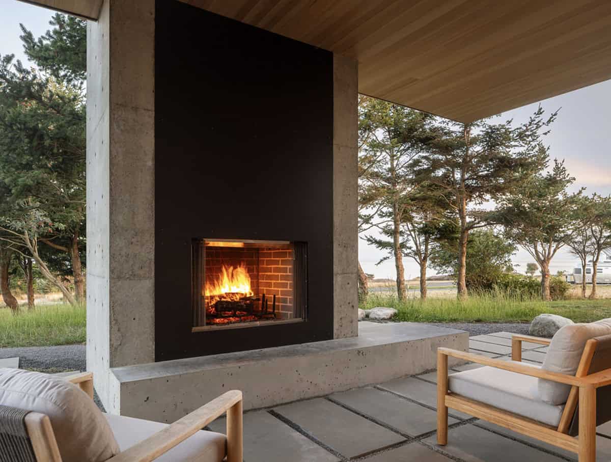 Outdoor fireplace with a roaring fire, flanked by concrete columns and teak lounge chairs at dusk