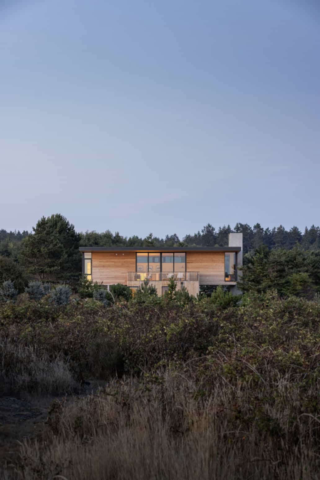 Distant dusk view of the wood-clad beach house glowing from within, set among trees and shrubland