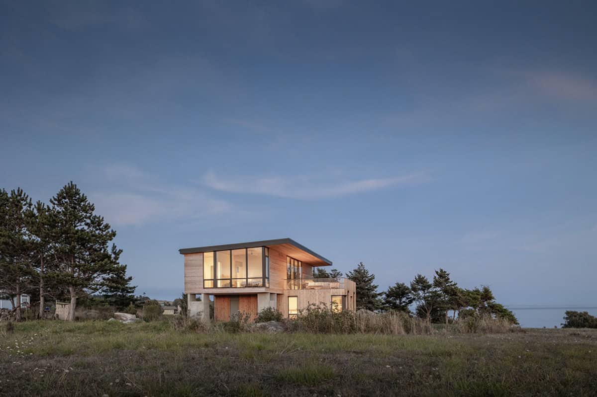 Twilight exterior of the two-story beach house with illuminated interiors, surrounded by grass and pines