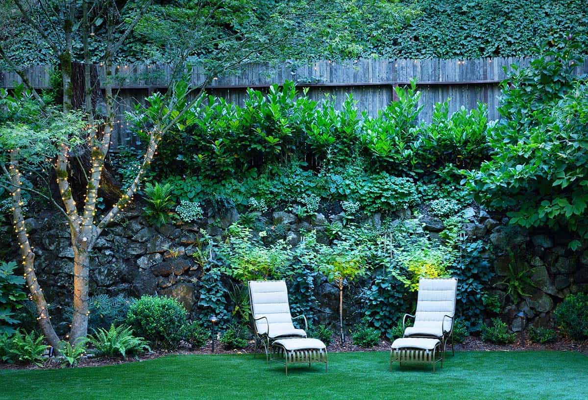 Two white lounge chairs on a lawn backed by a stone retaining wall draped in lush greenery and fairy lights
