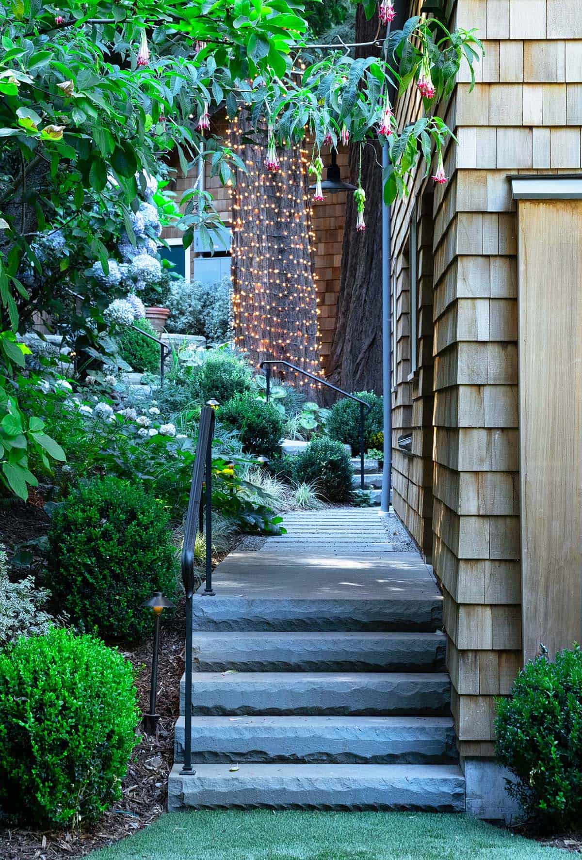 Stone steps with black iron railing alongside a cedar shingle wall, flanked by lush garden plantings and fairy lights