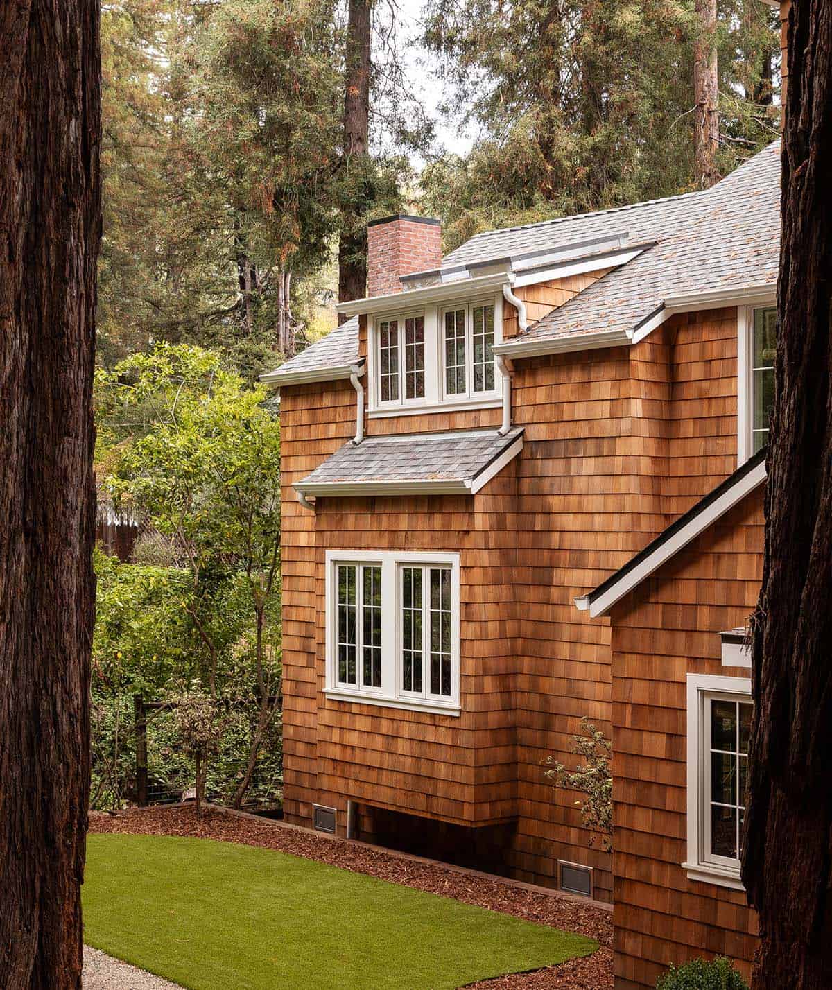 Close-up of cedar shingle facade with dormers, white-trimmed windows, and gray roof between redwood trees