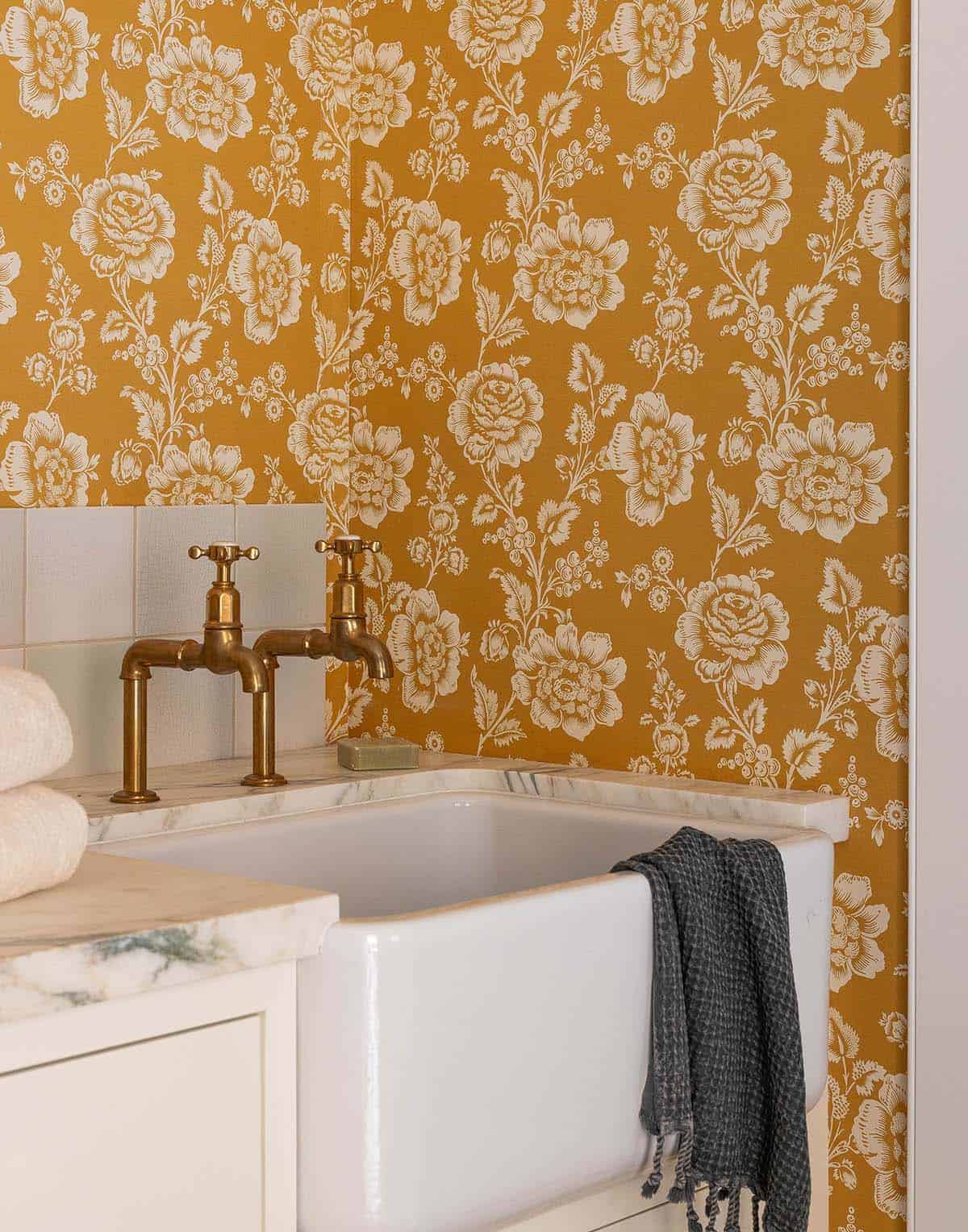 Farmhouse sink with unlacquered brass faucets against bold mustard yellow floral wallpaper