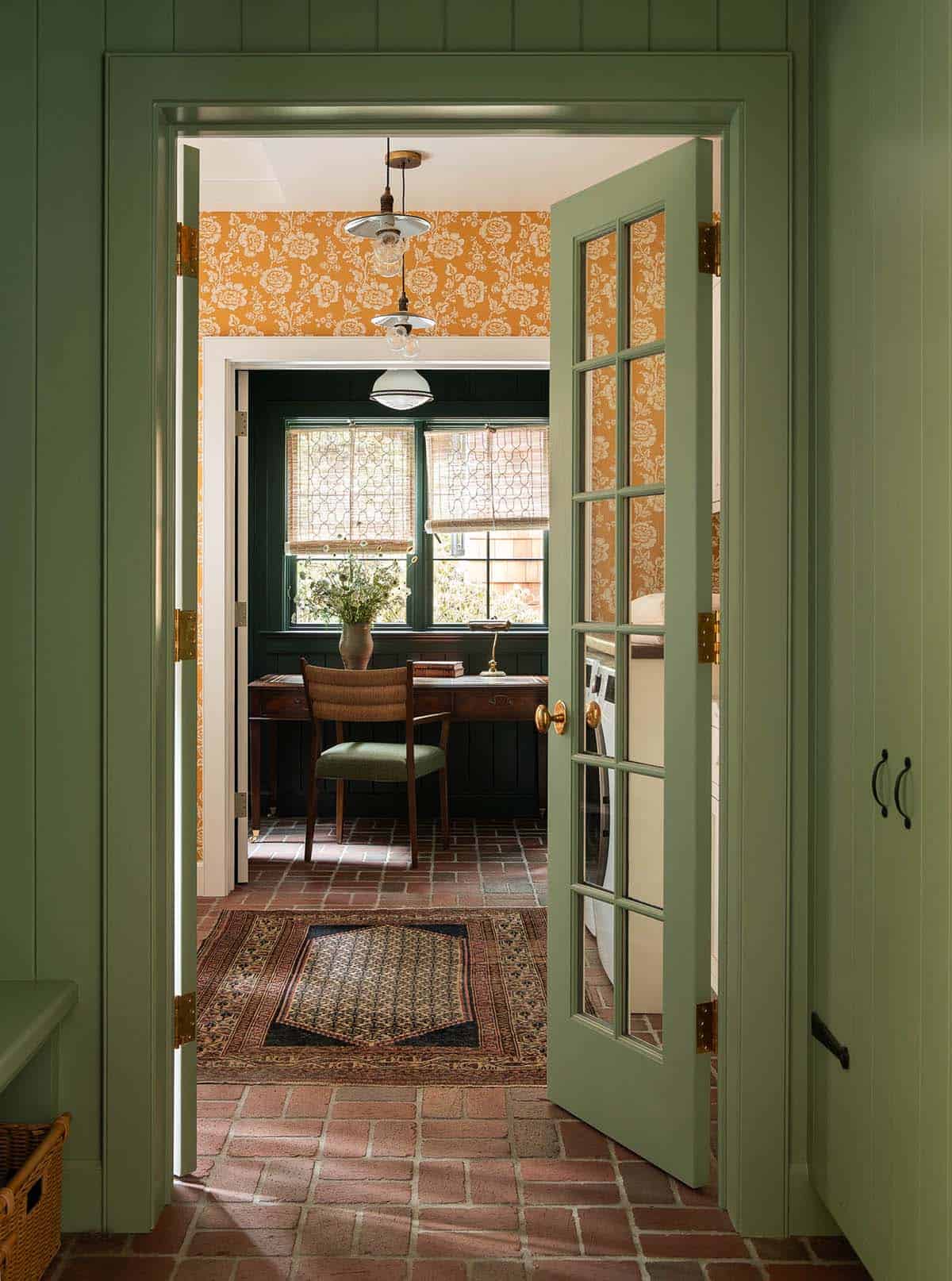 Green French doors opening into a home office with dark paneling, woven shades, and mustard floral wallpaper