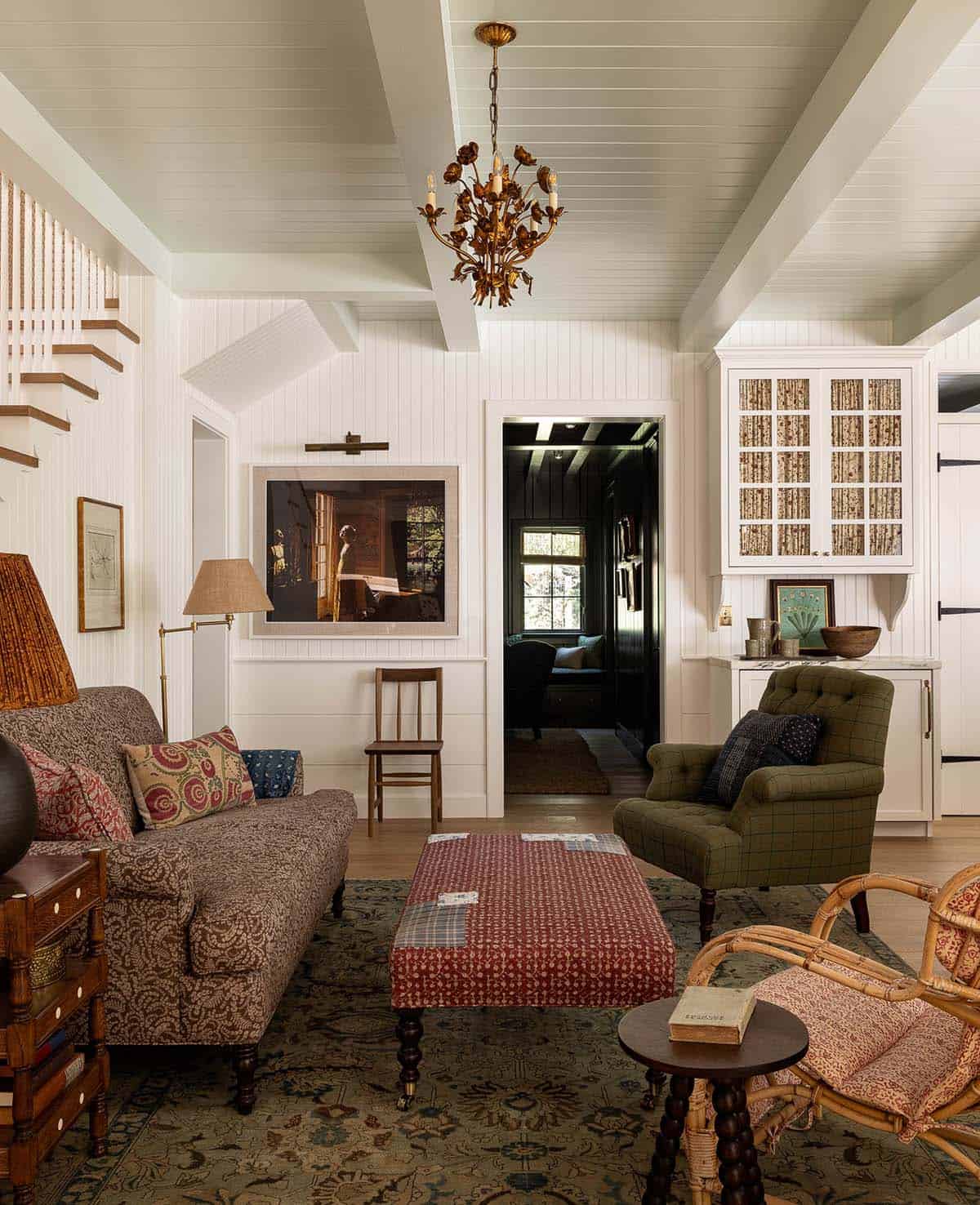 Bright living room with white beadboard walls, floral chandelier, layered vintage furnishings, and dark study beyond
