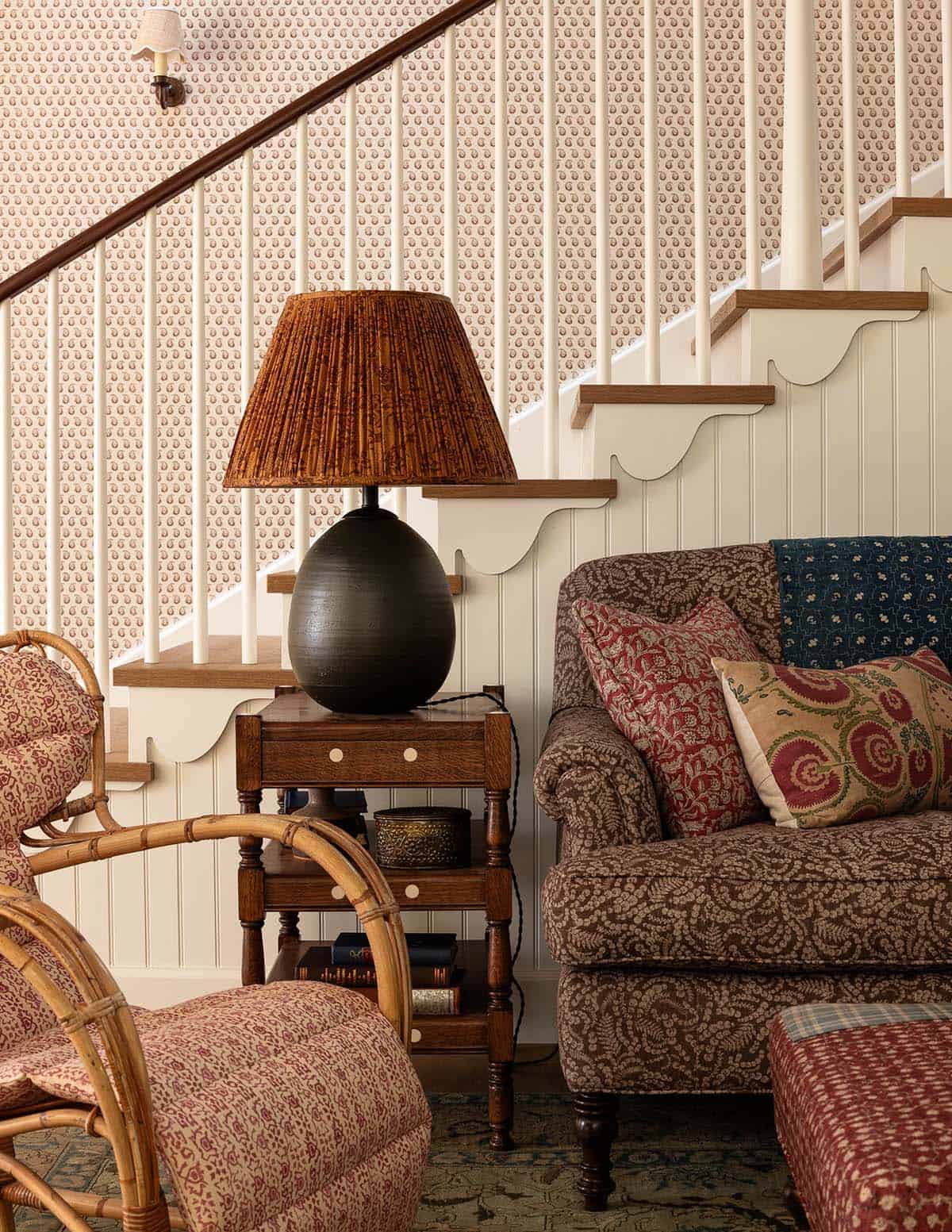 Staircase detail with printed wallpaper, scalloped baseboard trim, rattan chair, and a black ceramic table lamp
