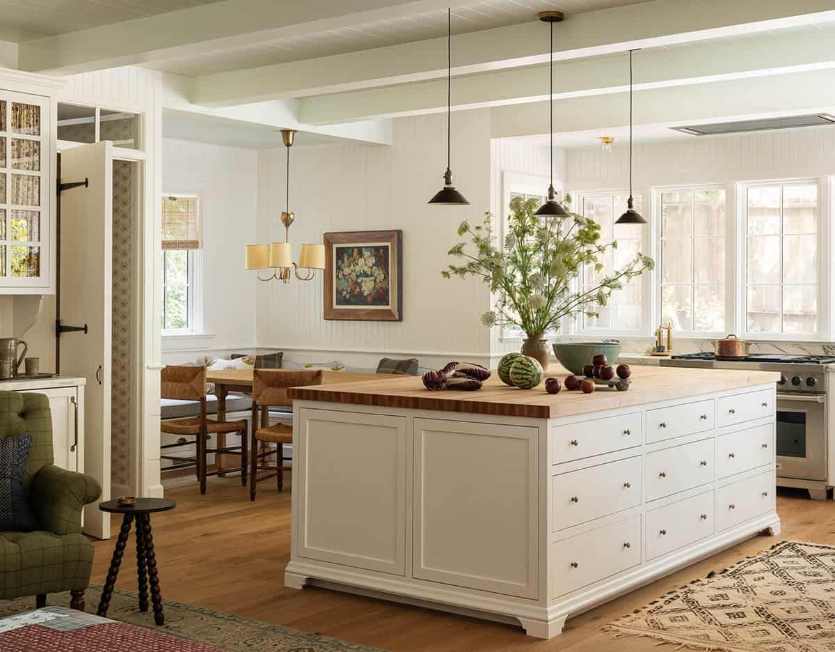 Large kitchen island with butcher block top, white cabinetry, brass hardware, and breakfast nook beyond