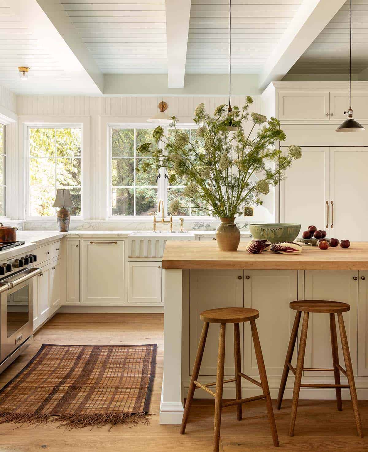 White kitchen with butcher block island, farmhouse sink, brass faucet, wood stools, and garden views