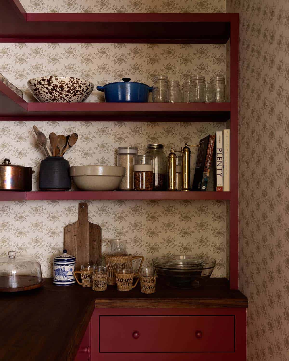 Pantry shelving with deep red painted shelves, floral wallpaper, and an eclectic mix of kitchenware and cookbooks