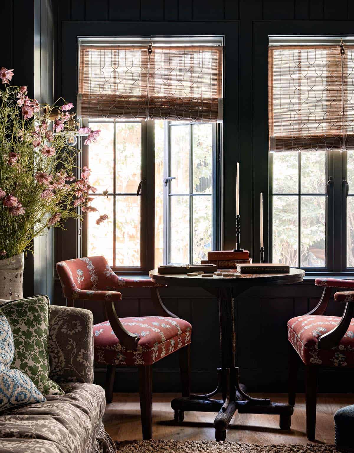Cozy dark-walled sitting area with two red floral chairs flanking a round pedestal table and bamboo shades
