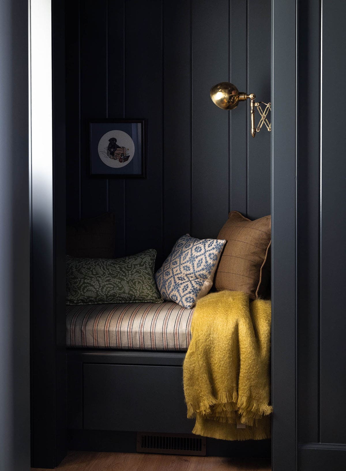 Built-in window seat nook with dark paneling, striped cushion, patterned pillows, and a brass articulating sconce