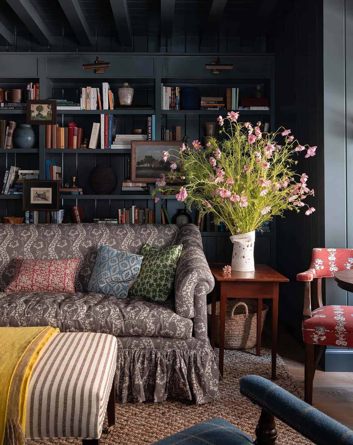 Dark library with patterned sofa, pink wildflower arrangement, built-in bookshelves, and red accent chair