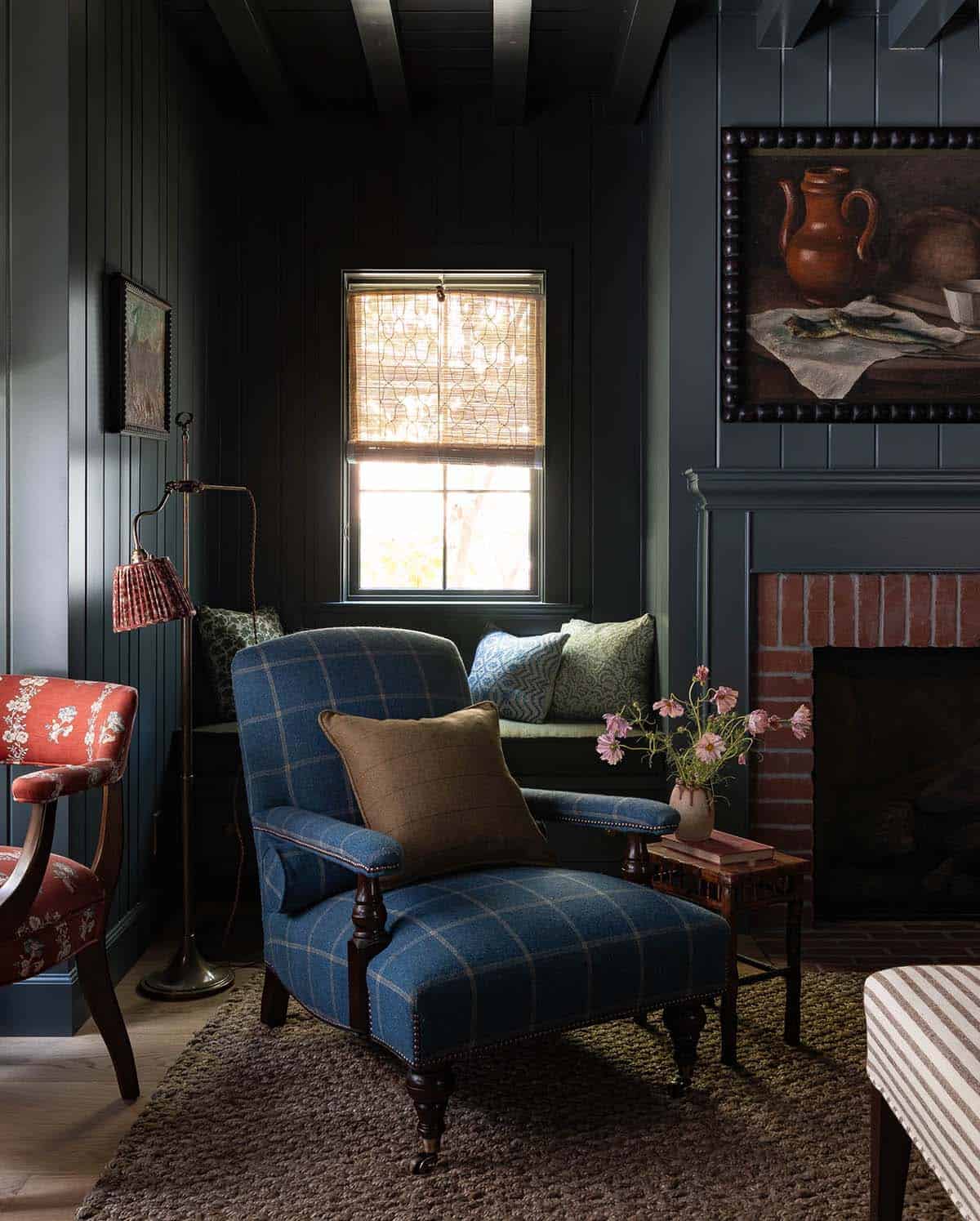 Dark paneled room with blue plaid armchair, brick fireplace, window seat, and still life painting above mantle
