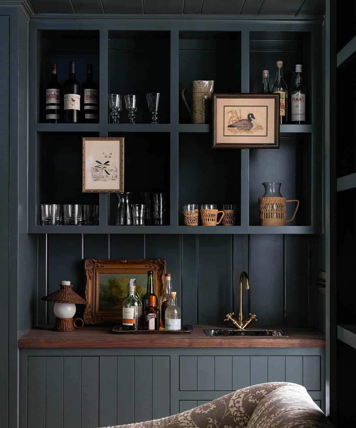 Dark teal built-in bar with open shelving, glassware, spirits bottles, wood countertop, and brass sink faucet