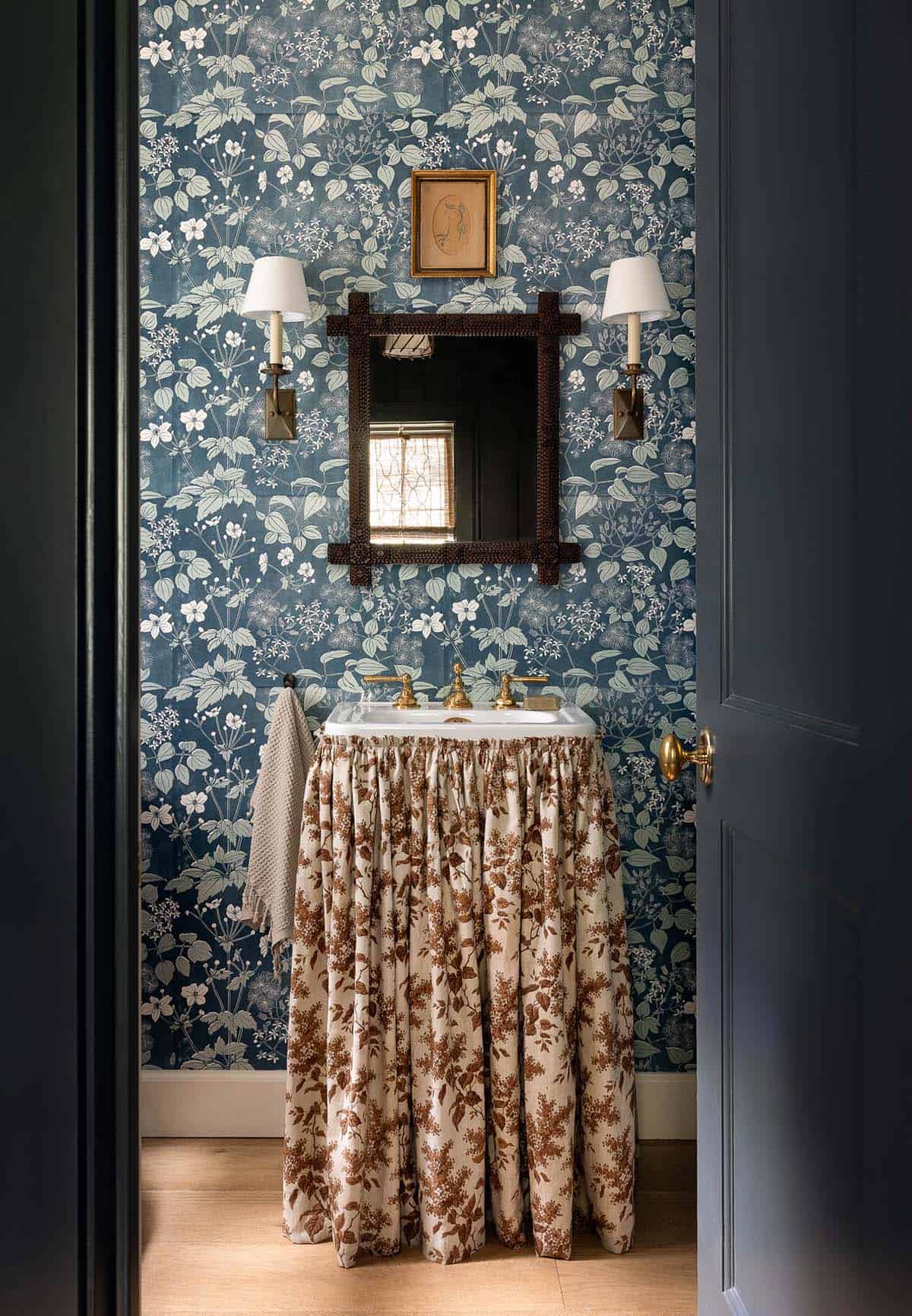 Powder room with bold navy botanical wallpaper, skirted vanity, dark wood mirror, and brass wall sconces