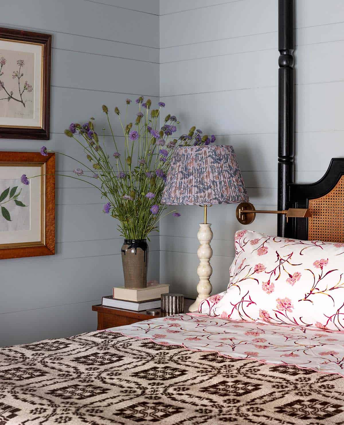 Bedroom nightstand detail with purple wildflowers, floral lamp shade, and pink floral pillowcases against blue-gray shiplap