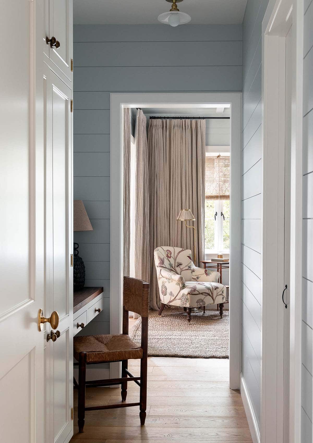 Blue-gray hallway with built-in desk nook leading to a sitting room with floral armchair and linen drapes