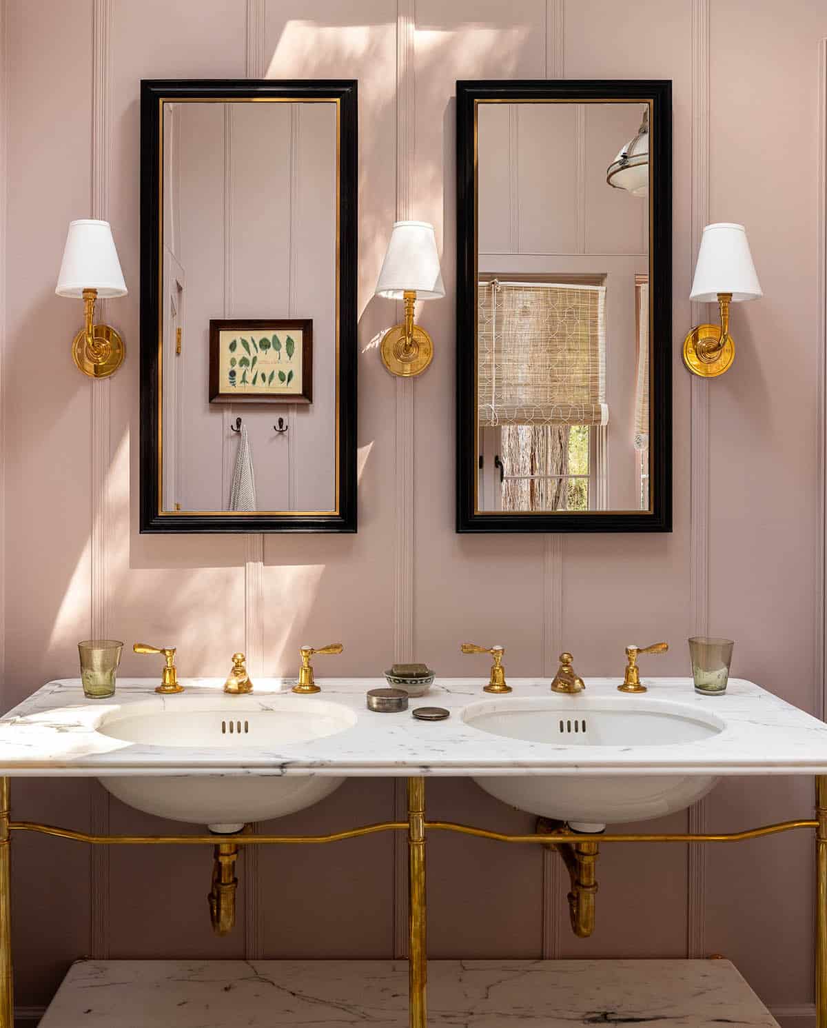 Double marble vanity with brass hardware, twin black-framed mirrors, and brass sconces on blush pink paneled walls