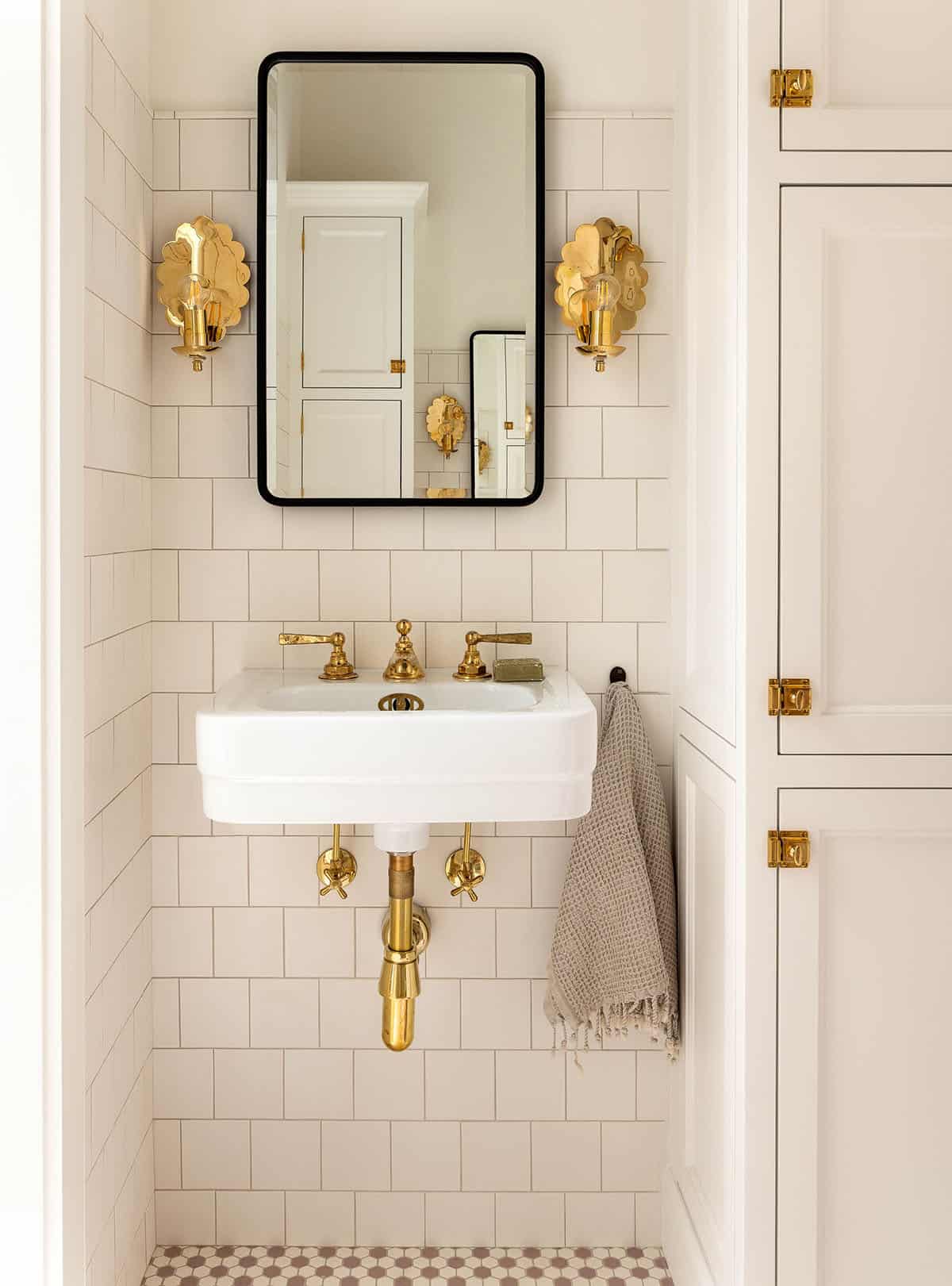 White-tiled bathroom with wall-mounted sink, brass fixtures, brass flower sconces, and black-framed mirror