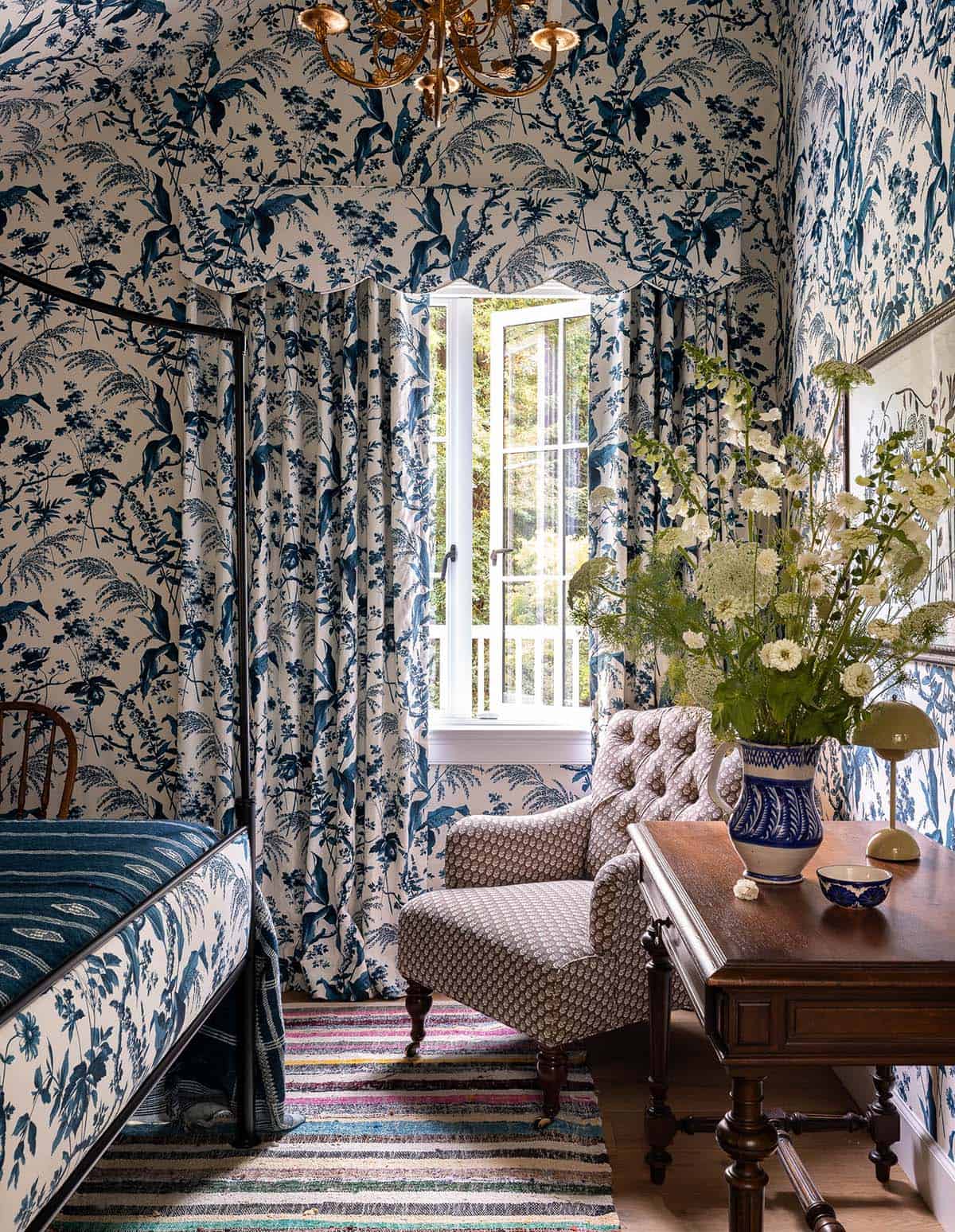 Blue toile bedroom corner with tufted armchair, matching curtains, open casement window, and white wildflower arrangement