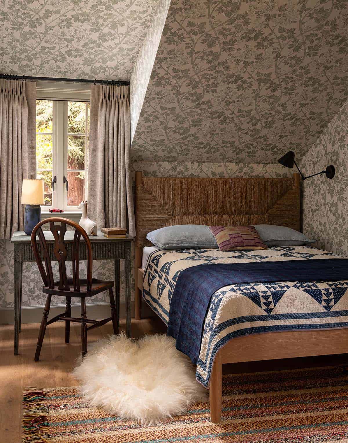Attic bedroom with all-over branch wallpaper, woven headboard, blue patchwork quilt, and small writing desk