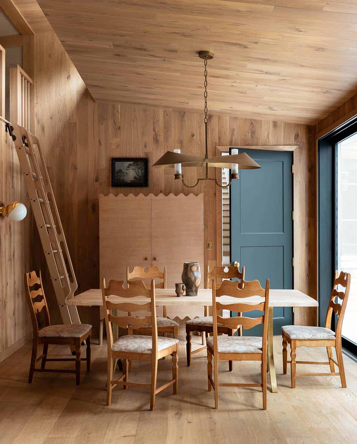 All-wood dining room with scallop-trimmed sideboard, carved oak chairs, brass chandelier, and teal painted door