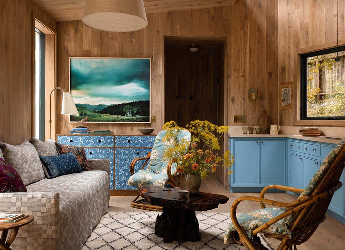 Wood-paneled guest cabin living room with blue patterned dresser, rattan chairs, live-edge coffee table, and landscape painting