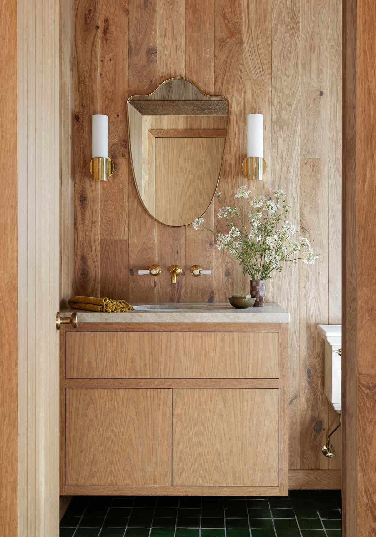 Wood-paneled bathroom vanity with shield-shaped mirror, brass wall-mounted faucet, stone countertop, and white candle sconces