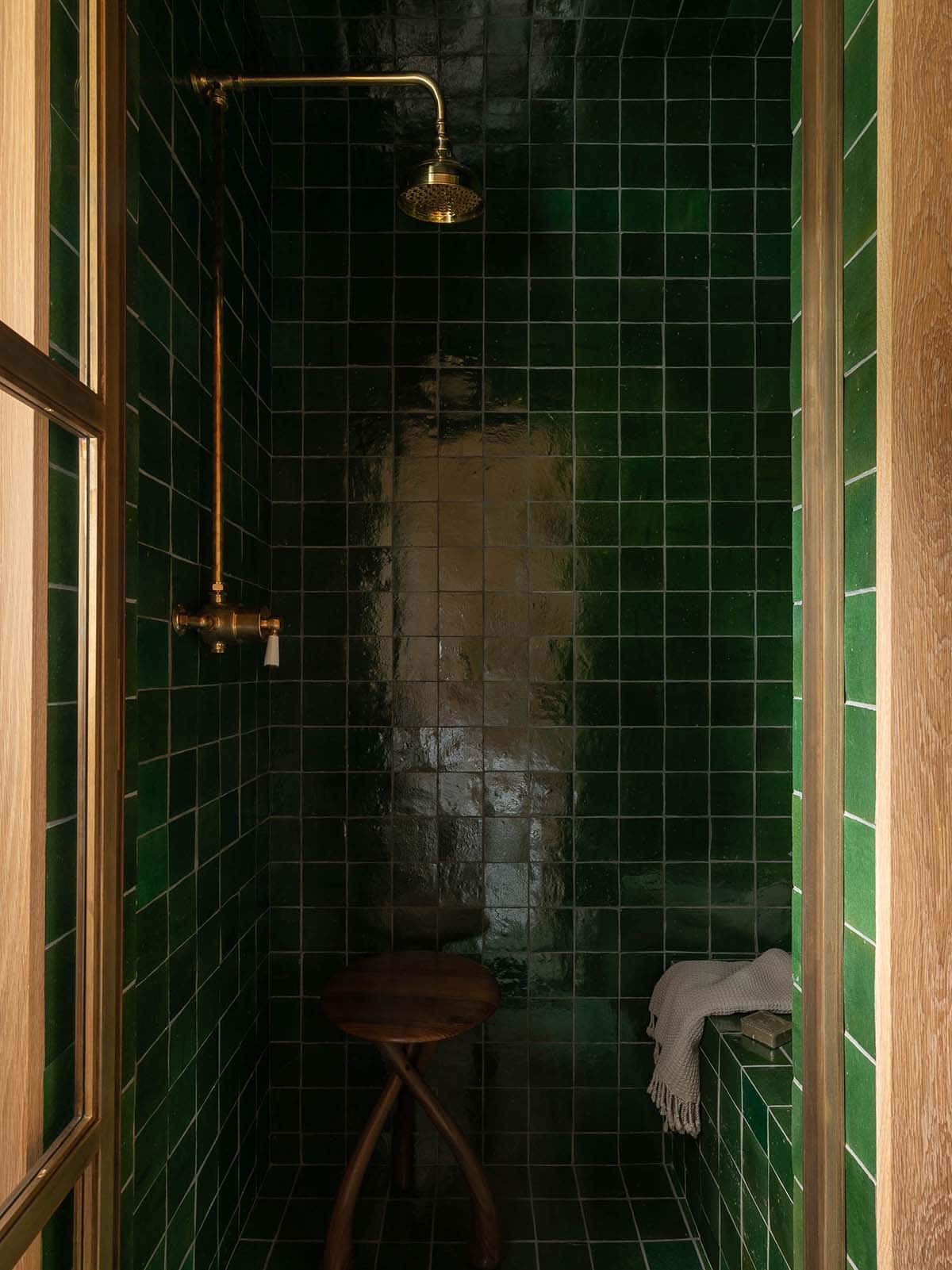 Deep green square-tiled shower with brass rainfall showerhead, wood stool, and oak door frame