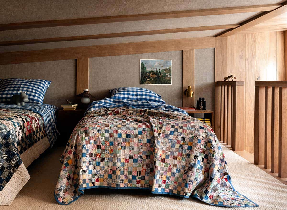 Children's bunk room with two beds, patchwork quilts, blue gingham pillows, and wood-paneled upholstered headboard wall