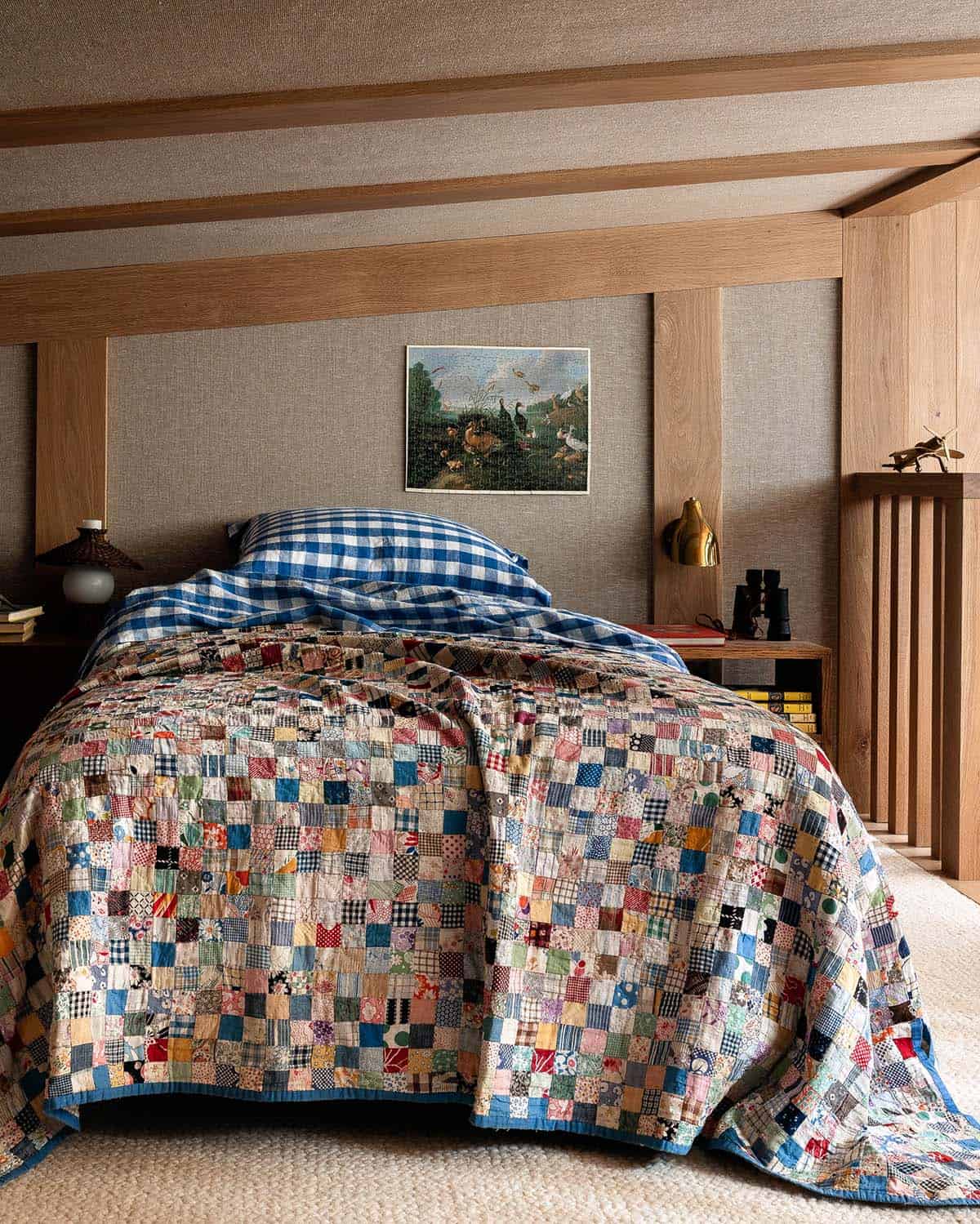 Close-up of bed with colorful patchwork quilt and blue gingham pillowcases against a linen and oak headboard wall